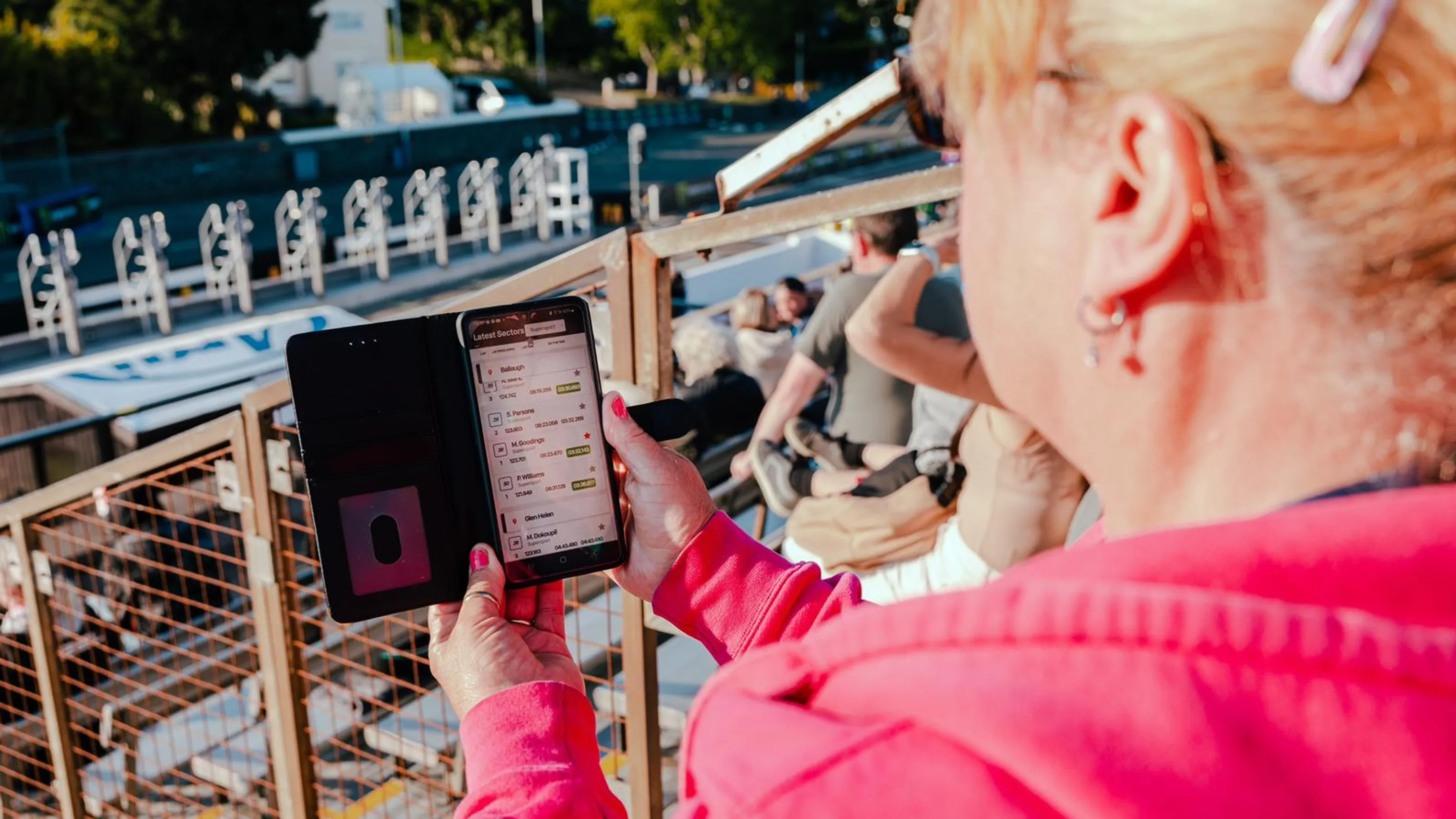 Woman in a pink hoodie holding a smartphone displaying the TT race timing app while watching from the grandstand or race enclosure